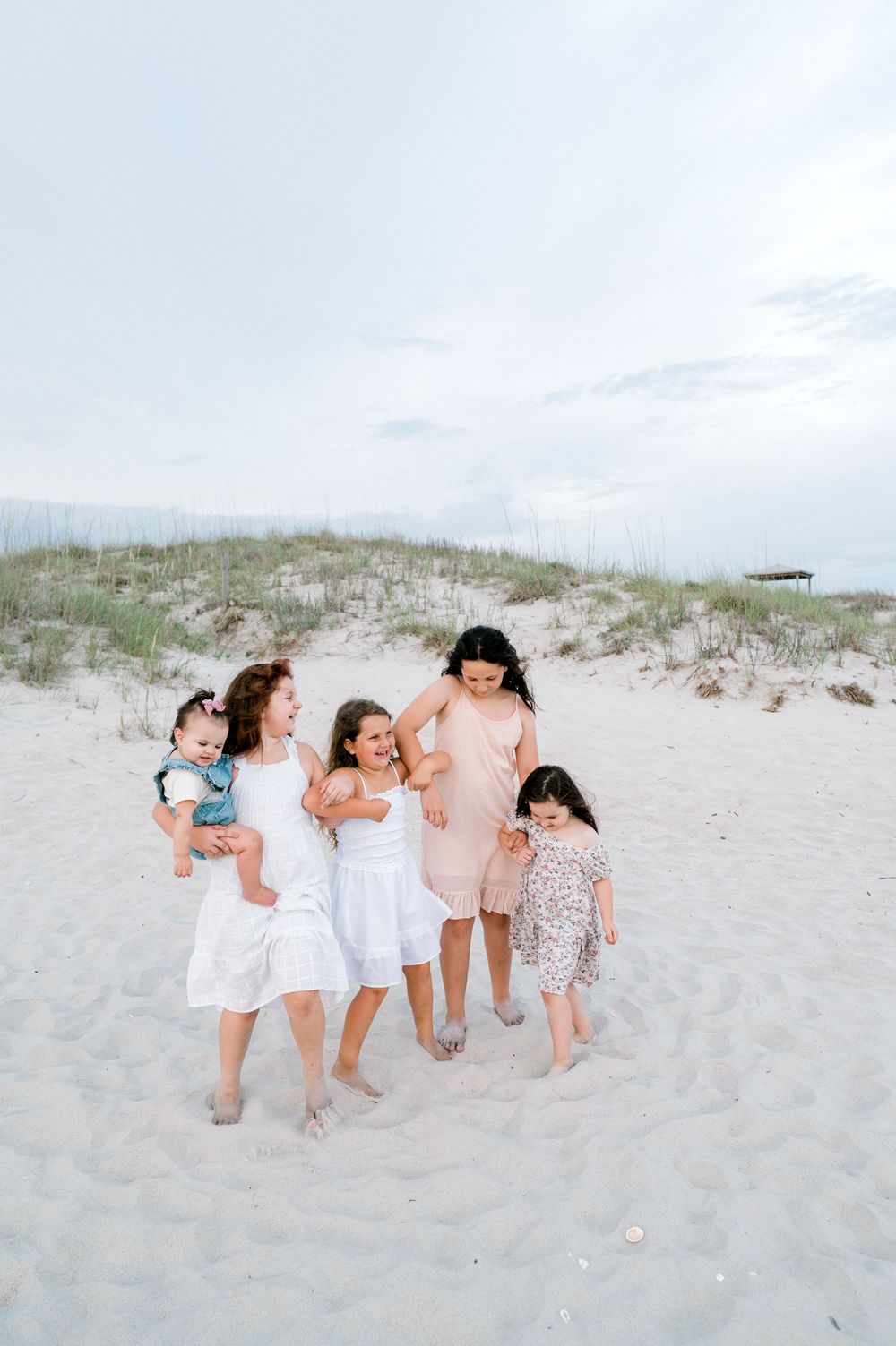 A joyful group of children and a woman walking together on a sandy beach near grass-covered dunes.