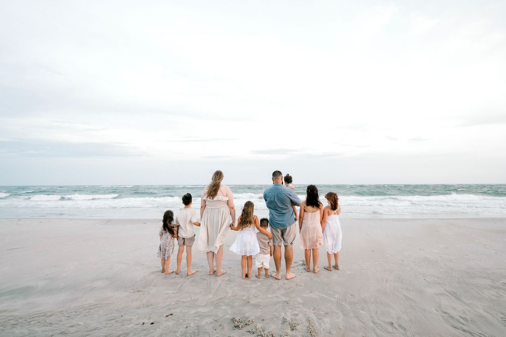 Large family holding hands on a sandy beach shore, facing the ocean waves under a soft cloudy sky.