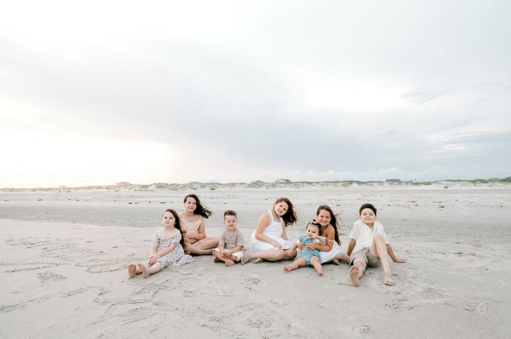 Family of seven sitting together on a sandy beach at sunset, dressed in white and neutral tones.