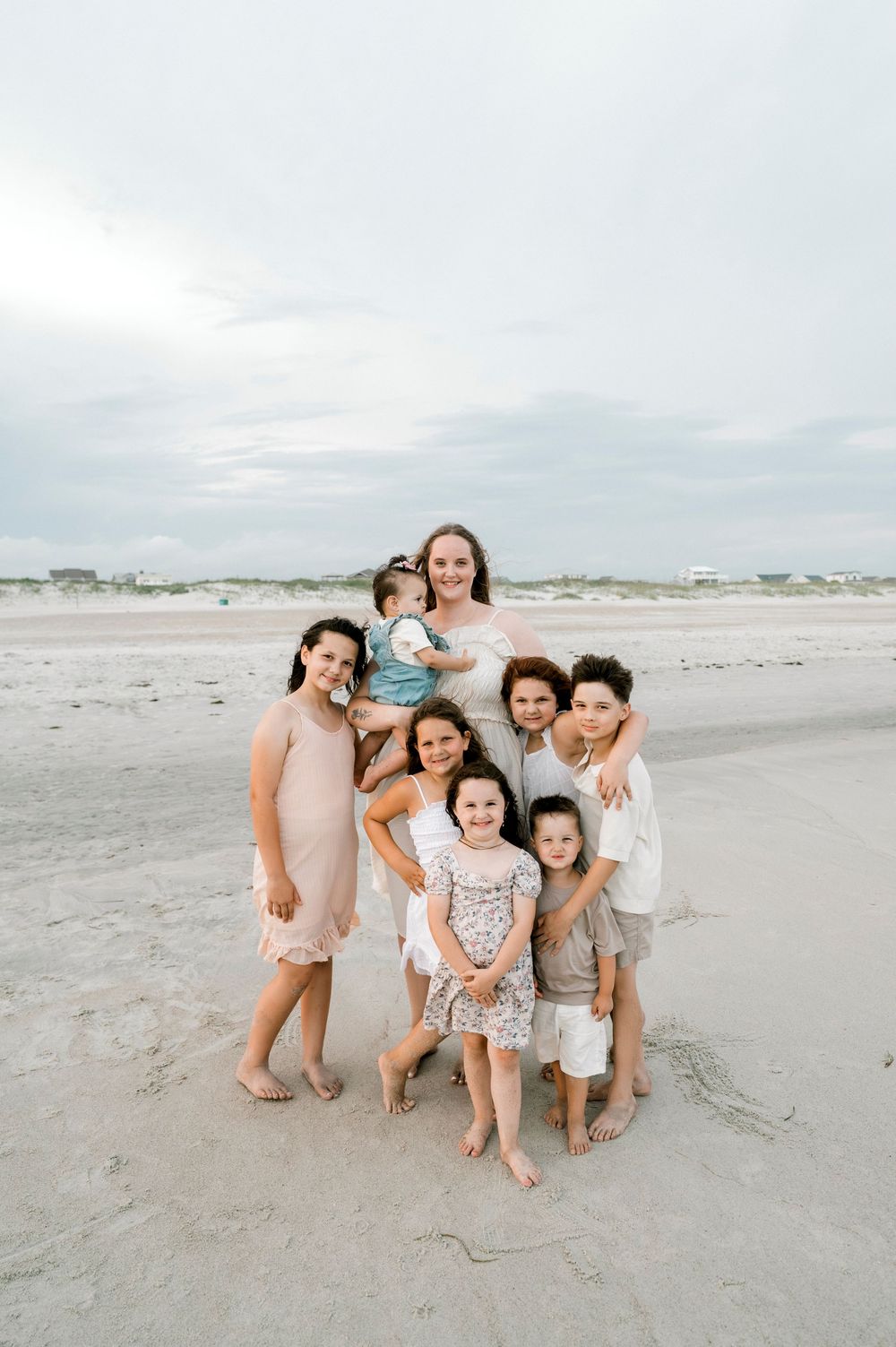 A woman and six children pose together smiling on a sandy beach under a cloudy sky.