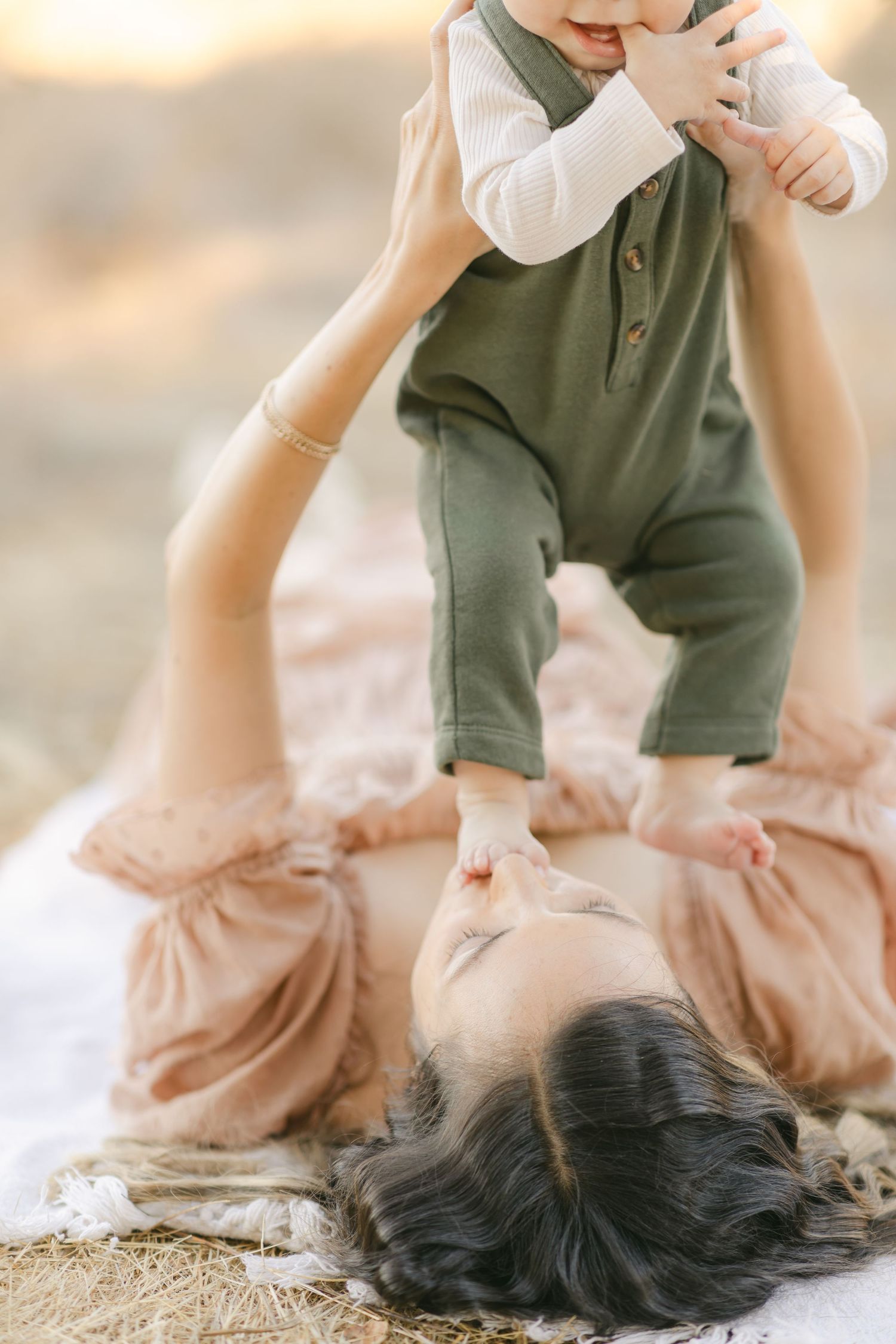 A mother lying on a blanket outdoors, lifting her baby dressed in a green romper in a warm, golden-lit setting.