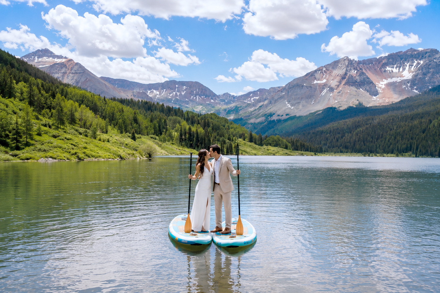 Couple in wedding attire standing on a paddleboard on a serene mountain lake with lush green trees and rocky peaks.
