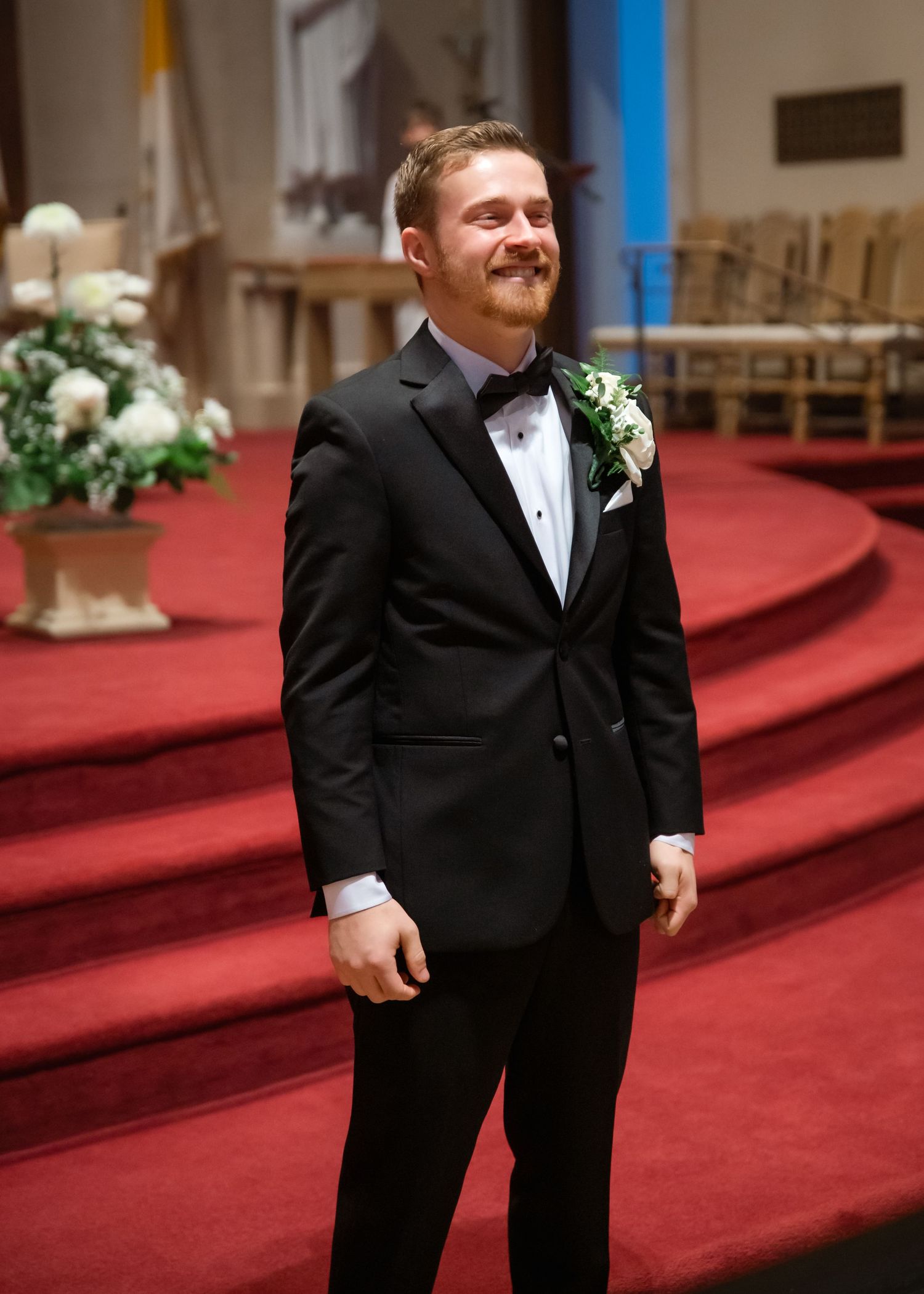 Smiling groom in black tuxedo with boutonniere standing at church altar with red carpet and floral arrangements.