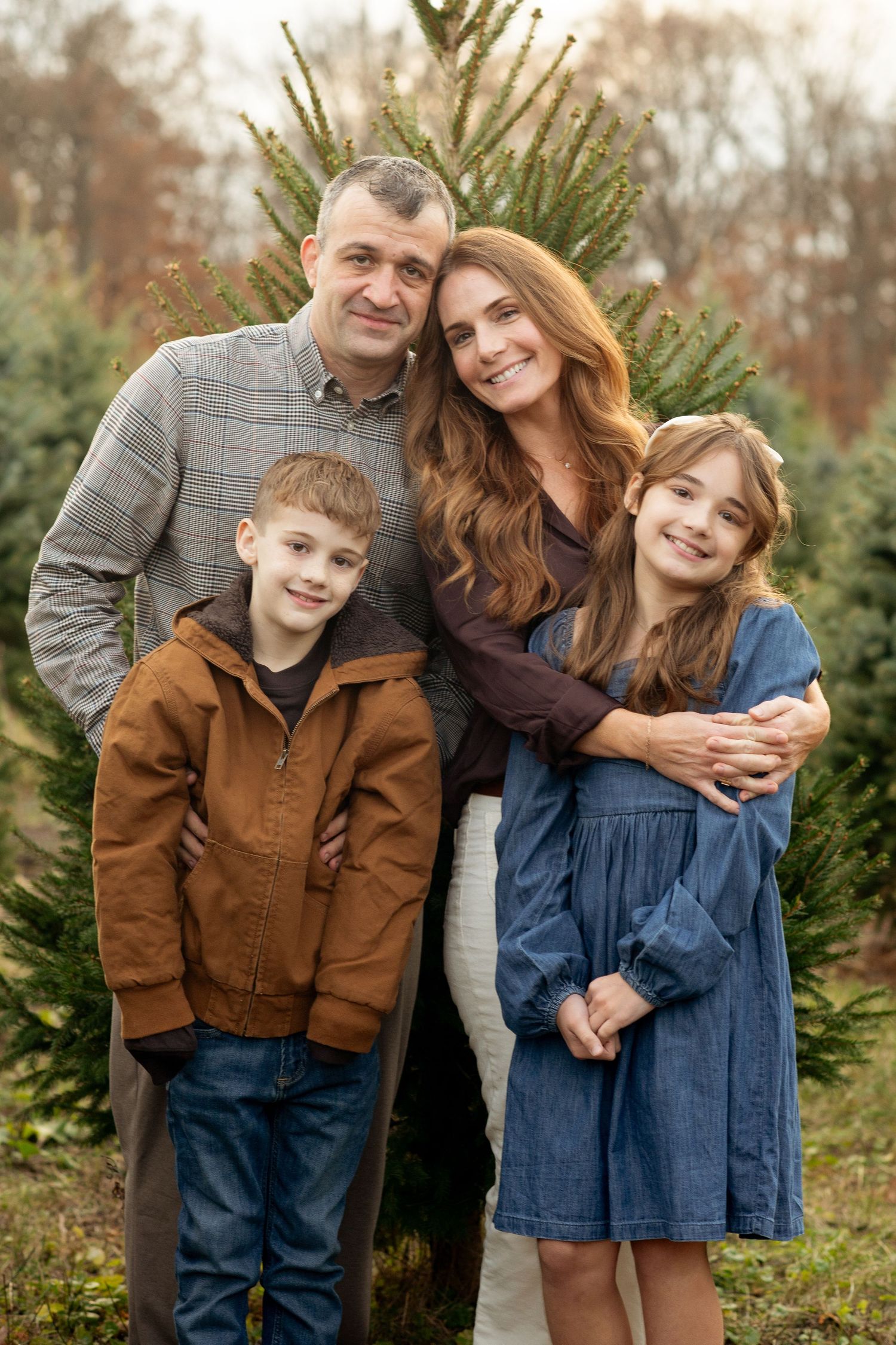 Family of four posing at a Christmas tree farm, parents with two children smiling outdoors among green pine trees.