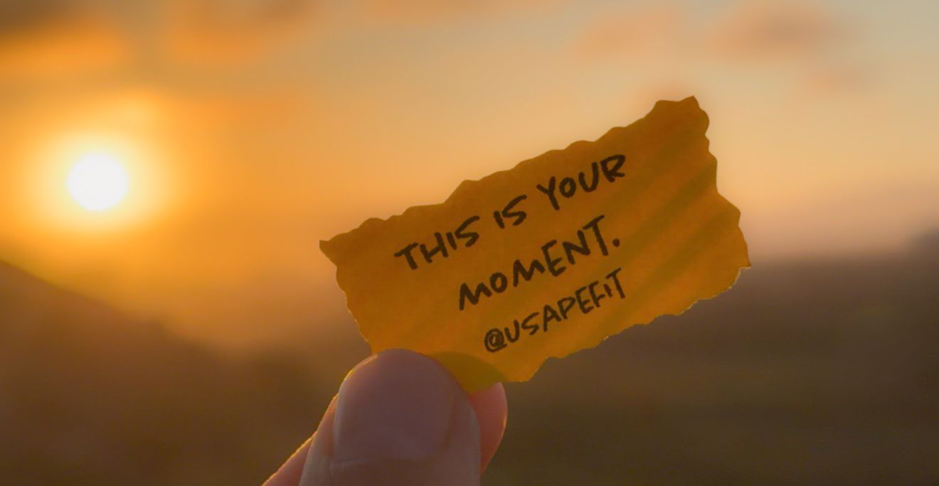 Hand holding a torn paper note reading This Is Your Moment at golden sunset with mountain backdrop.