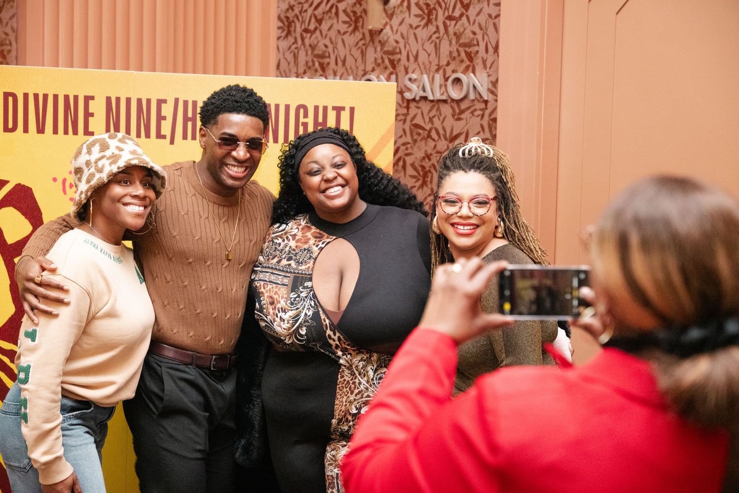 Four people pose for a photo at a Divine Nine event, smiling while someone captures the moment on a smartphone.
