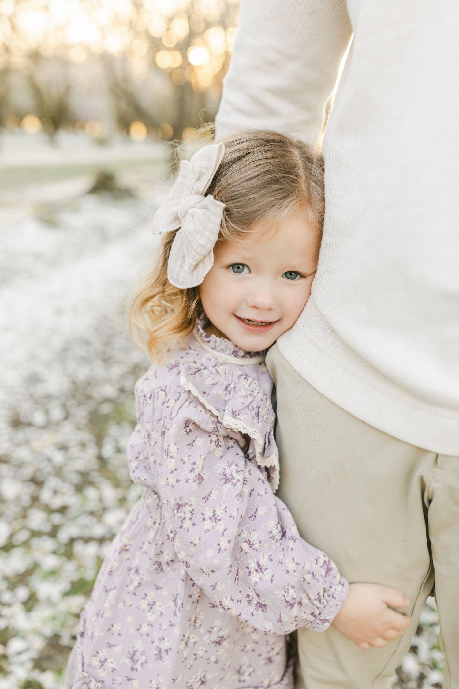 A smiling toddler girl in a floral dress and bow hugs an adults leg outdoors during golden hour.