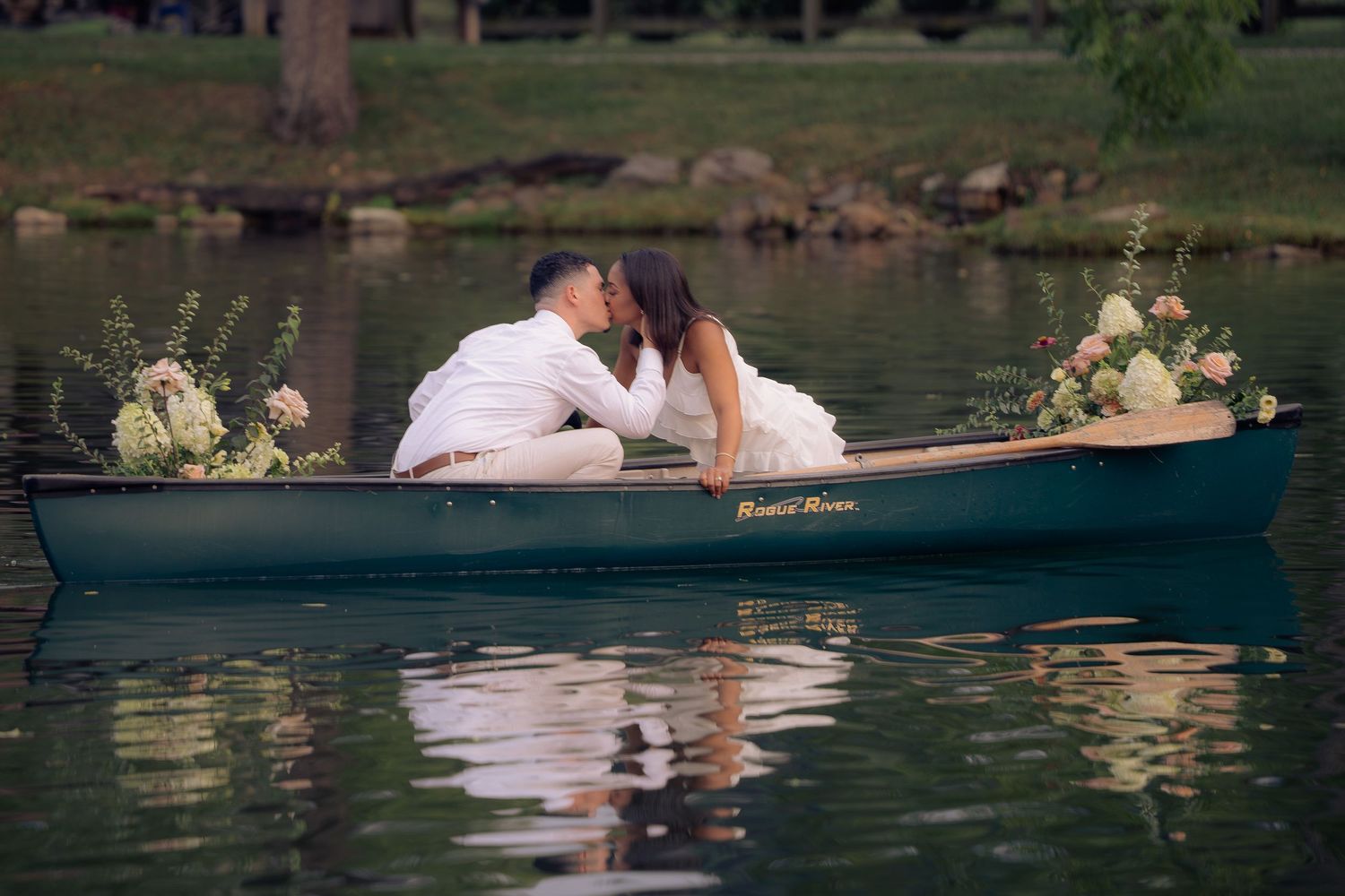 Newlywed couple kissing in a green canoe on a calm lake, surrounded by lush floral arrangements.