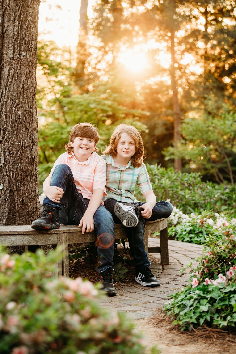 Two smiling boys sitting on a wooden bench in a sunlit garden surrounded by blooming flowers.