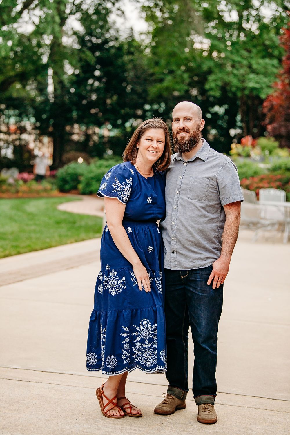 Smiling couple posing together on a garden pathway, woman in blue dress, man in gray shirt.