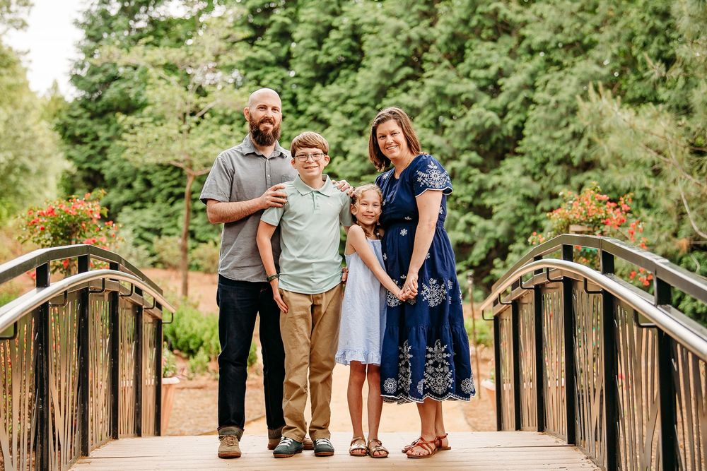 Family of four standing on a wooden bridge surrounded by green trees and flowers.