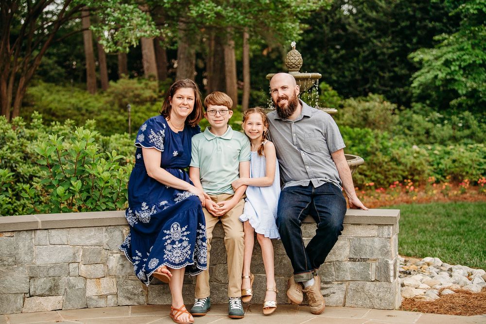 Family of four sitting and smiling together on a stone wall in a garden setting.