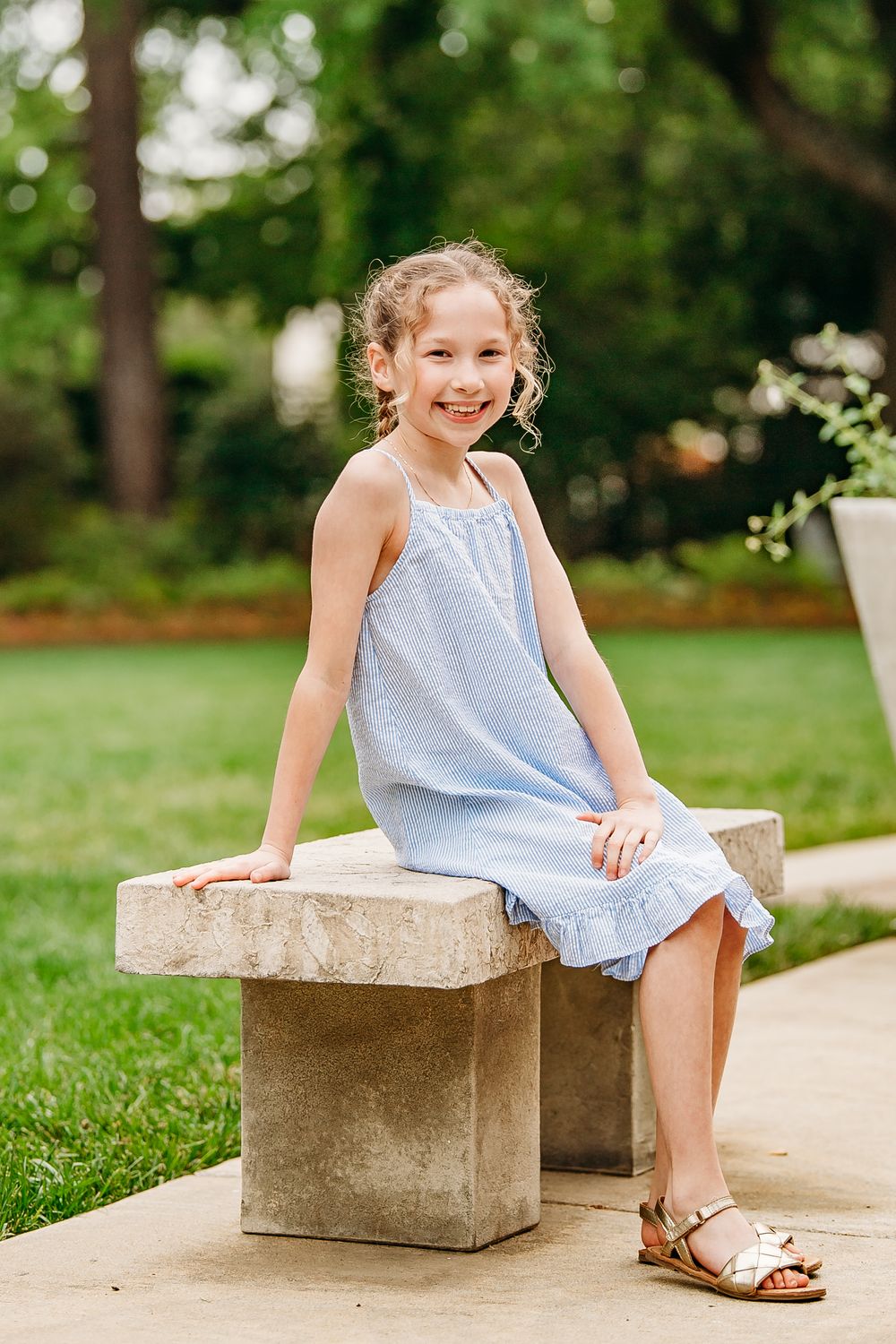 Smiling young girl in a light blue dress sitting on a stone bench in a park.