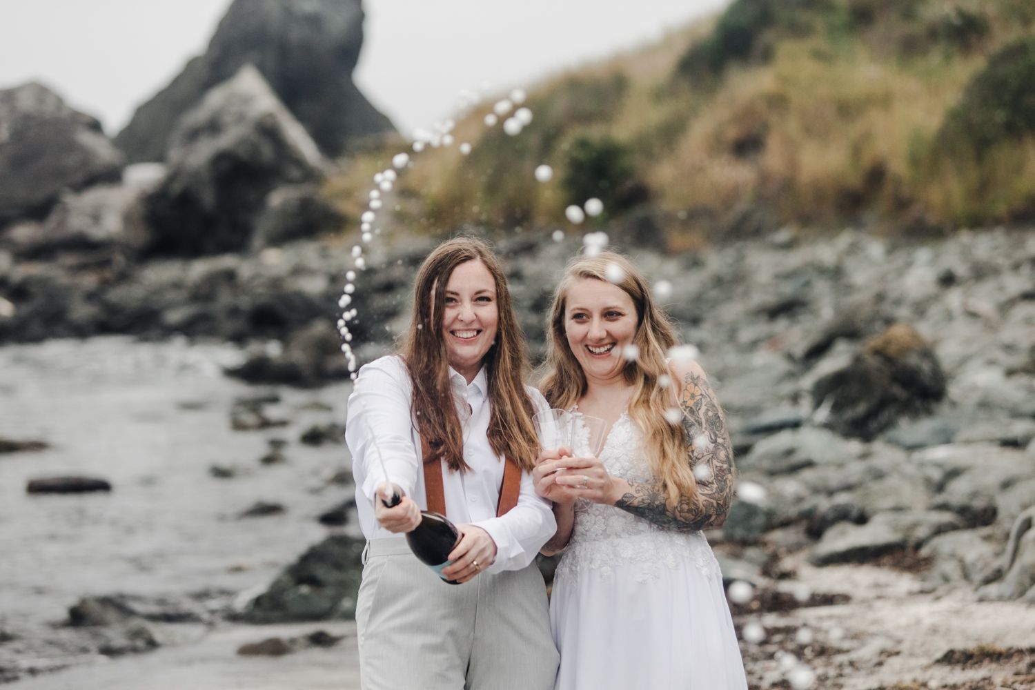 Couple pops champagne and sprays towards the camera with the rocky Oregon Coast in the background
