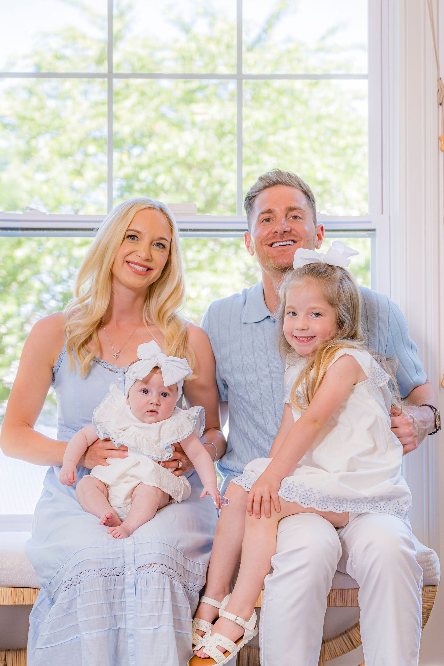 A smiling family of four poses indoors, with parents holding a baby and young girl in white and blue outfits by a bright window.