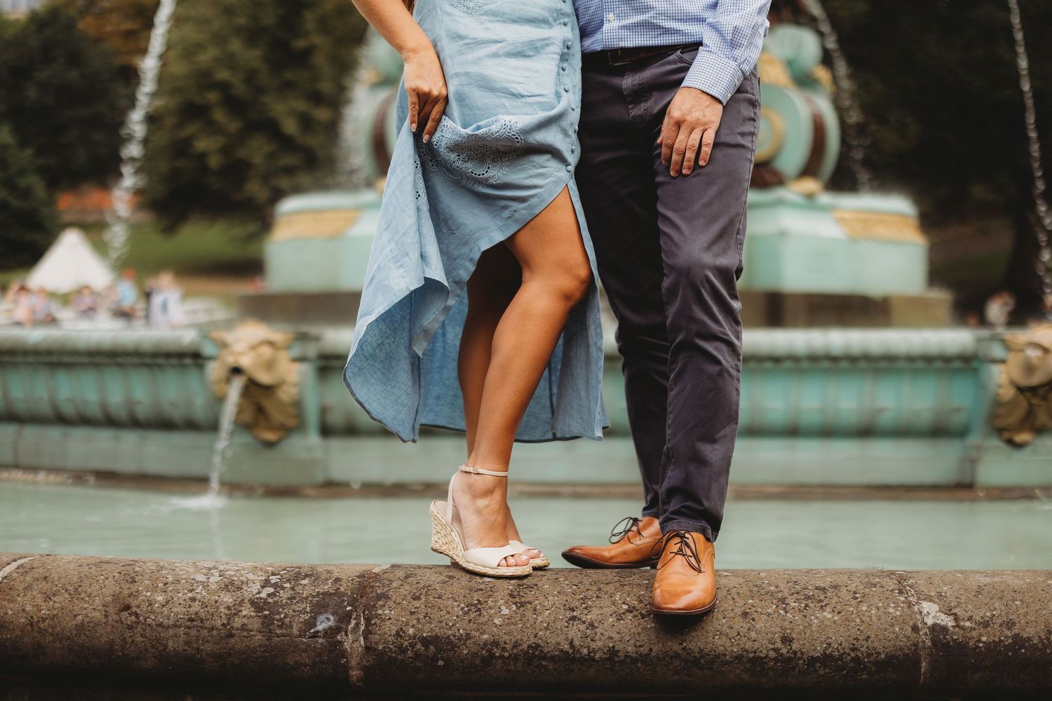 Couple standing by a decorative fountain, wearing blue dress and brown shoes, sharing a romantic moment outdoors.