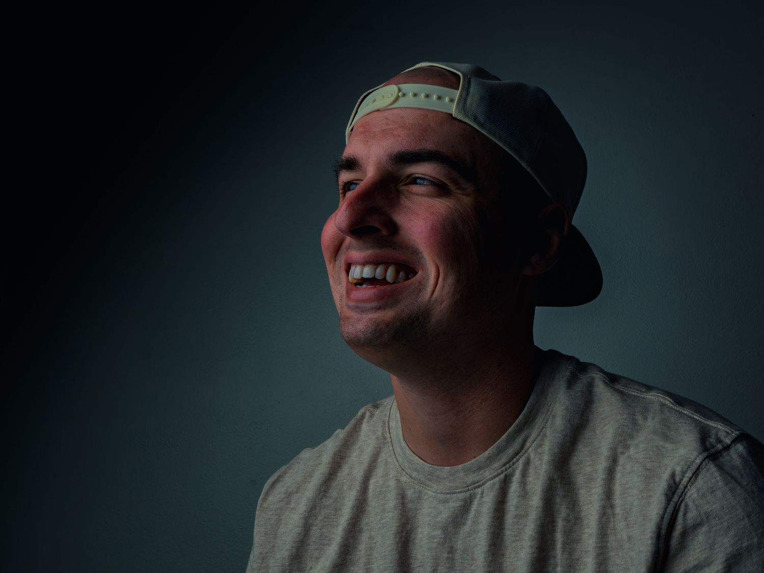 A man in a backward cap and gray tee laughs while looking off to the side, warm light washing across his face against a dark teal wall