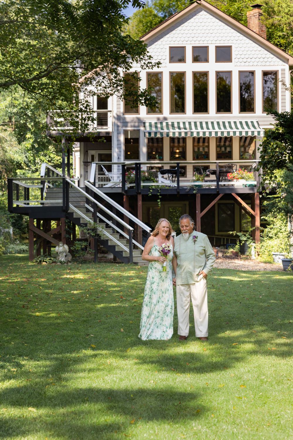 Bride in floral dress and older woman pose on green lawn before elegant two-story home with deck.