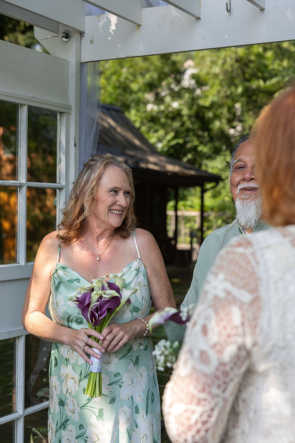 Woman in floral dress holds purple calla lily bouquet, smiling at bride during outdoor wedding ceremony.