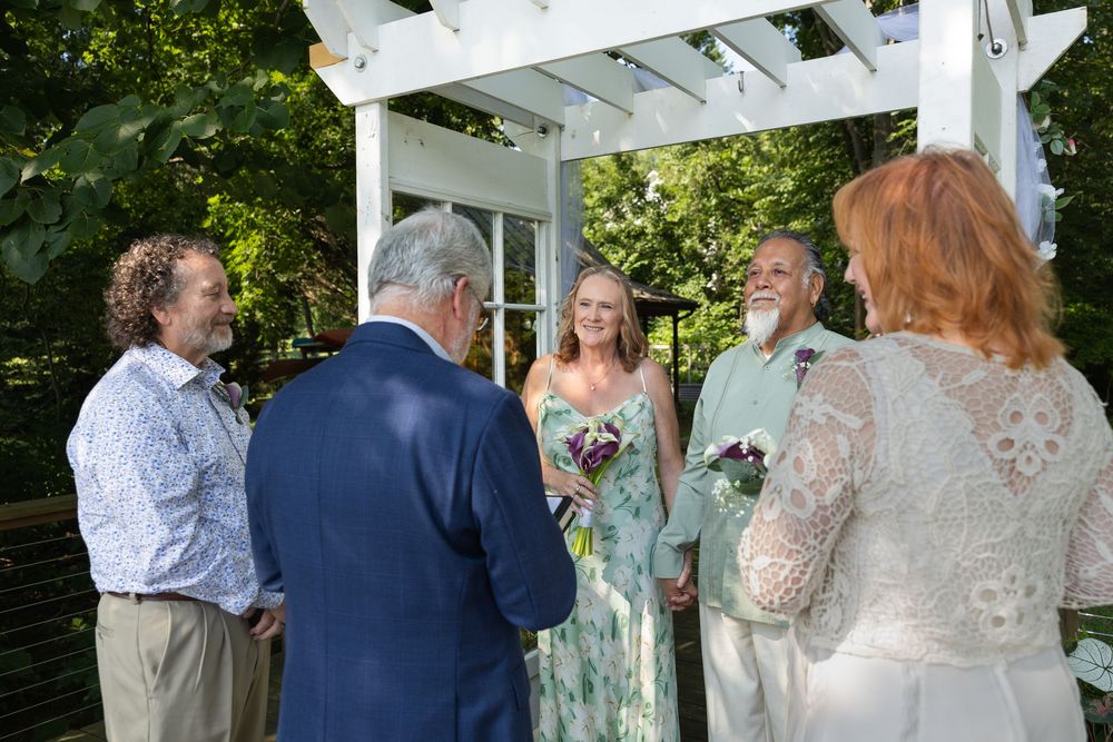 Small outdoor wedding ceremony under a white pergola with guests gathered around a smiling bride holding flowers.