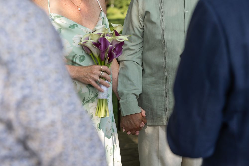 Couple holding hands at wedding ceremony, bride holding purple calla lily bouquet in elegant outdoor setting.