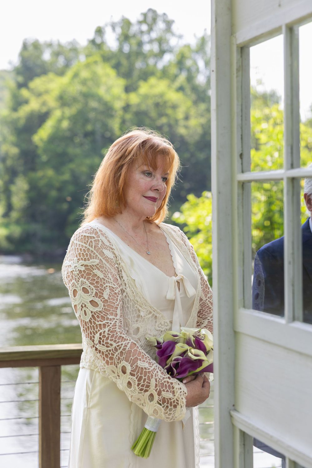 Elegant bride in lace jacket holding purple tulip bouquet on decorated porch near water.