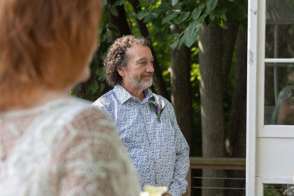 Man in floral shirt smiling warmly outdoors at a wedding ceremony near trees and white doors.