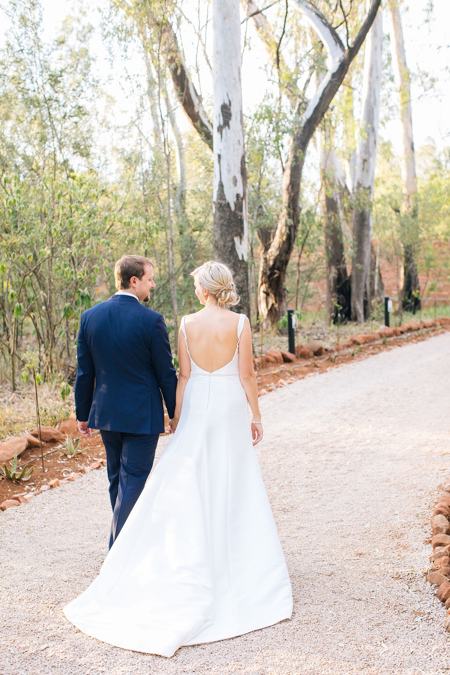 Bride and groom walking hand in hand down a tree-lined path, bride wearing a backless white gown and groom in navy suit.