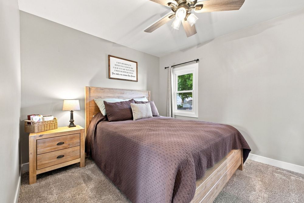 Cozy bedroom with queen bed, brown bedding, wooden nightstand, ceiling fan, and carpeted floors in a bright, neutral-toned room.