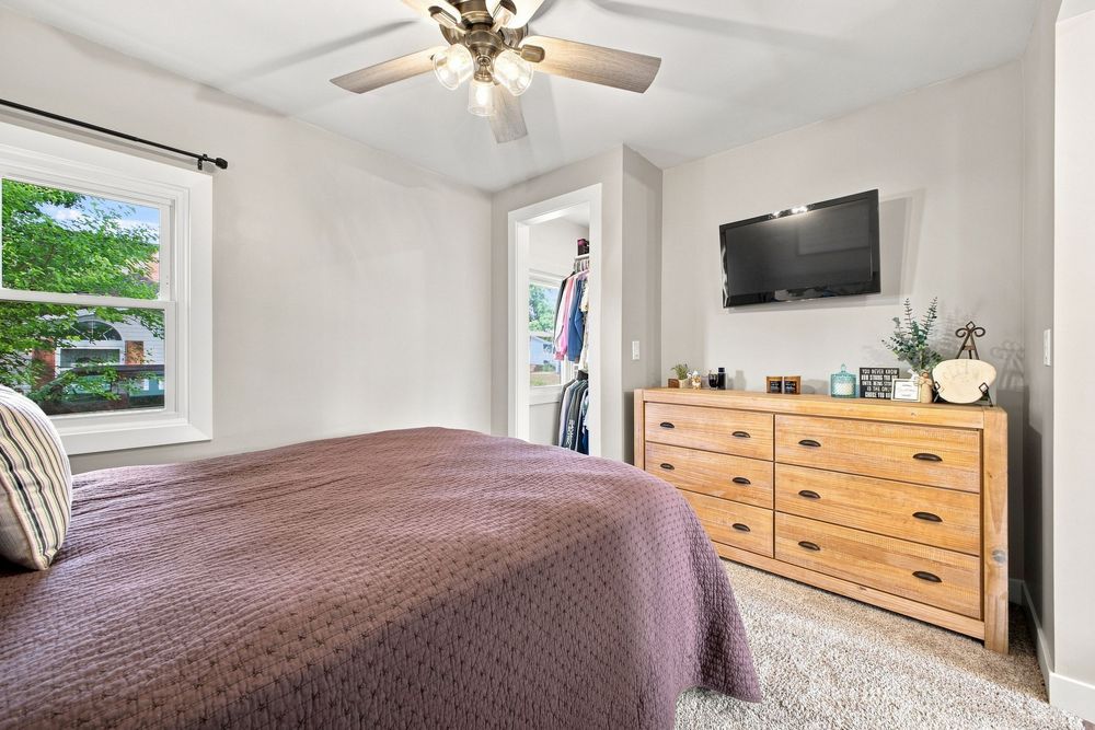 Cozy bedroom with purple bedspread, wooden dresser, wall-mounted TV, ceiling fan, and walk-in closet.