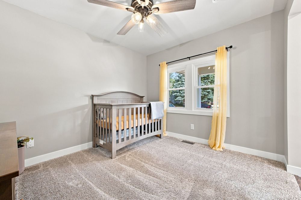 Bright nursery with gray crib, beige carpet, ceiling fan, and yellow curtained window in a neutral-toned room.