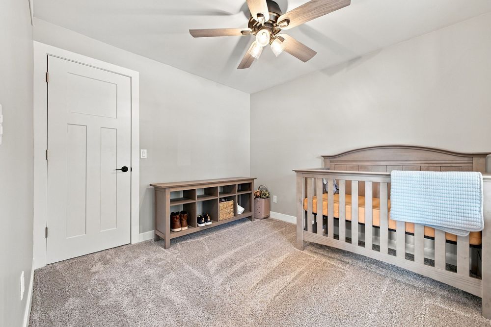 Nursery room with gray crib, ceiling fan, carpet flooring, storage bench, and white door in a neutral-toned space.