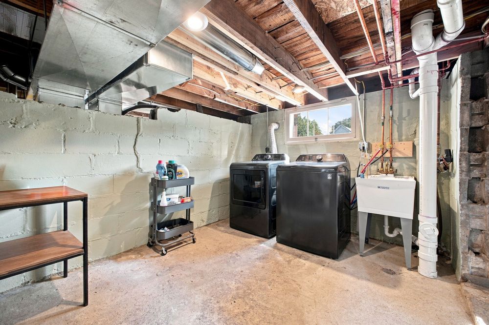 Unfinished basement laundry room with black washer and dryer, utility sink, exposed ceiling joists, and concrete walls.