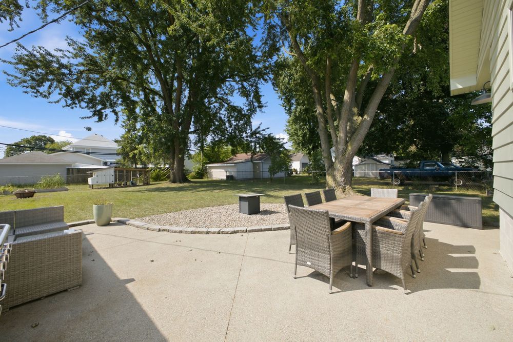 Outdoor patio with wicker dining set on concrete, surrounded by lush trees and a gravel fire pit area in a spacious backyard.