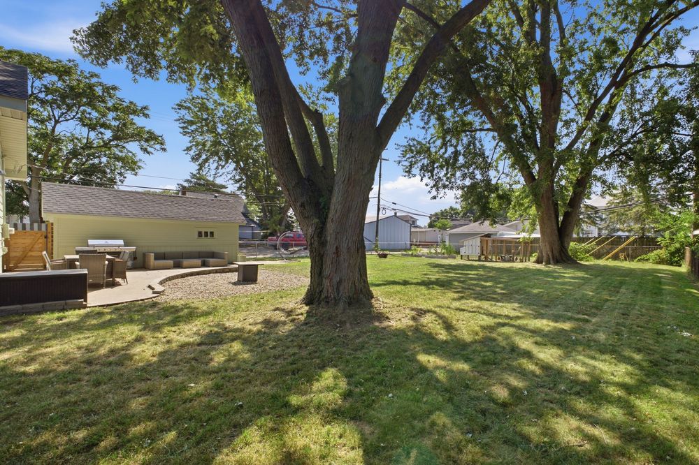 Spacious backyard with lush green lawn, large mature trees, patio area, and neighboring homes visible on a sunny day.