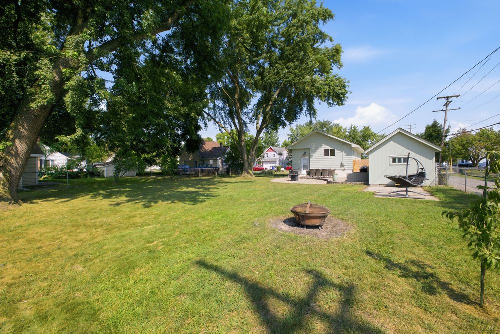 Spacious backyard with lush green lawn, mature trees, fire pit, and neighboring homes visible under a clear blue sky.
