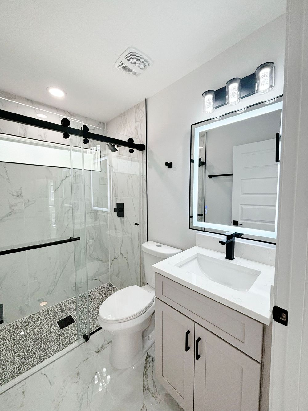 Modern bathroom with marble floors, glass shower, LED mirror, white vanity, and black hardware accents.