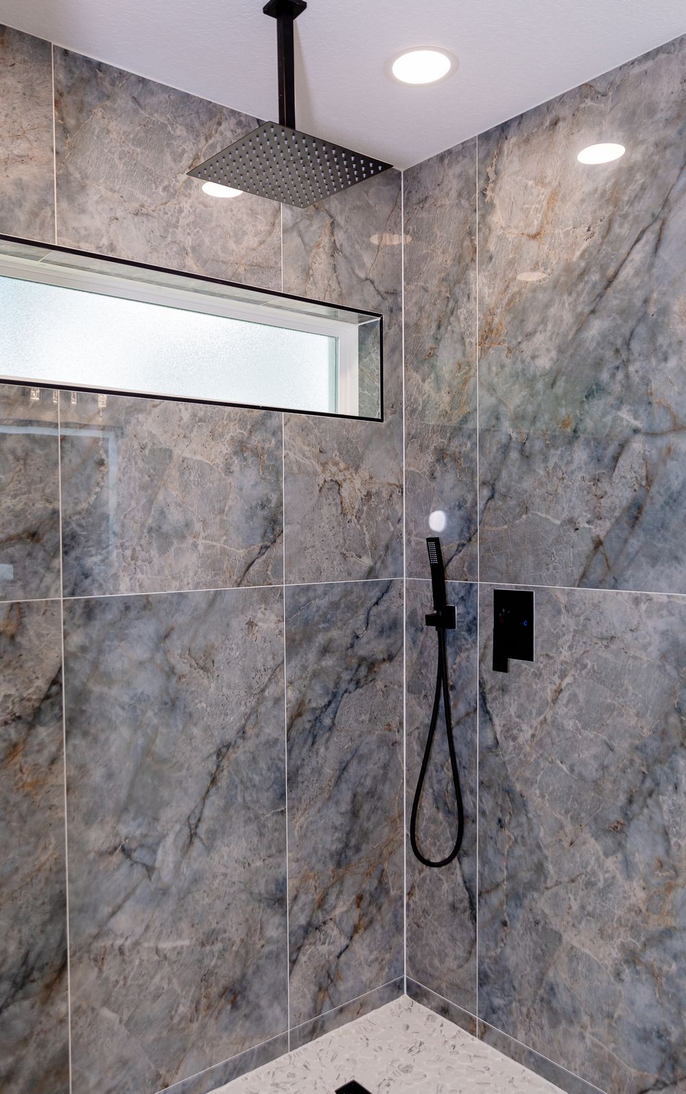 Modern walk-in shower with large marble-look tiles, black rainfall showerhead, handheld sprayer, and frosted window.