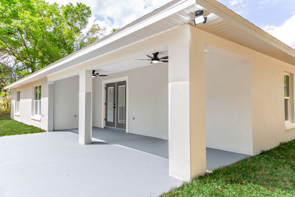 Modern home exterior featuring a covered patio with ceiling fans, French doors, and a spacious concrete driveway surrounded by lush trees.