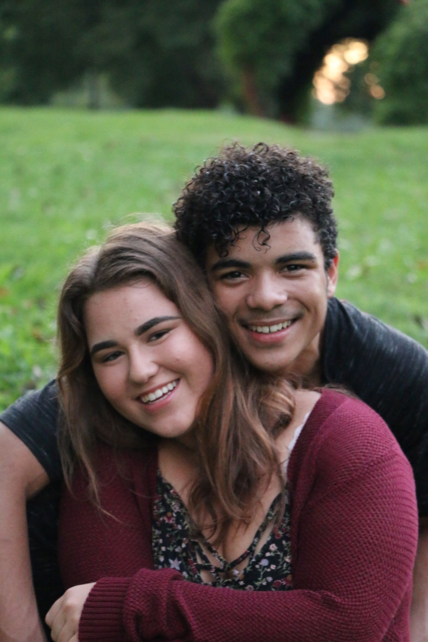 Two smiling teenagers embrace outdoors on green grass, girl with long brown hair and boy with curly hair wearing maroon sweaters.