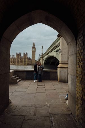 Couple posing under a stone arch with Big Ben and Westminster Bridge in the background on a cloudy day.