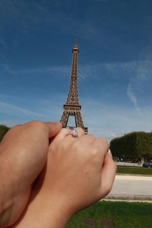 Couple holding hands with engagement ring in front of the Eiffel Tower in Paris on a sunny day.