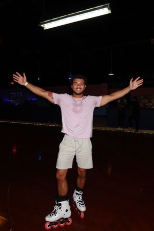 Young man in pink shirt and white shorts wearing inline skates, arms outstretched joyfully at a roller skating rink.