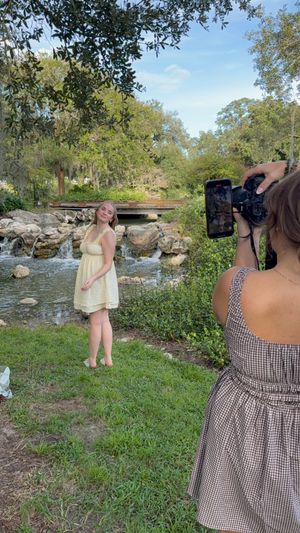 A photographer captures a young woman in a yellow sundress posing by a scenic creek with rocks and lush greenery.