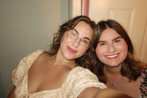 Two young women smiling together for a selfie, one wearing glasses and a floral dress.