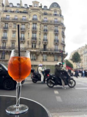 Aperol Spritz cocktail in a wine glass with a straw on a cafe table, with Parisian architecture and motorcycles in the background.