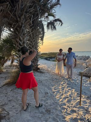 Photographer capturing a couple posing on a sandy beach at sunset near a palm tree with golden sky backdrop.