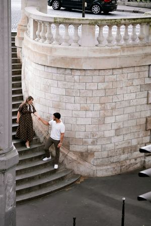 Couple holding hands on ornate stone staircase beside curved balustrade wall in European city.