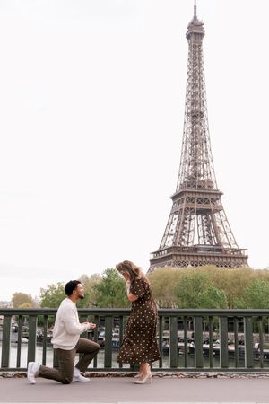 Man proposes to woman on one knee on a bridge with the Eiffel Tower in Paris, France in the background.