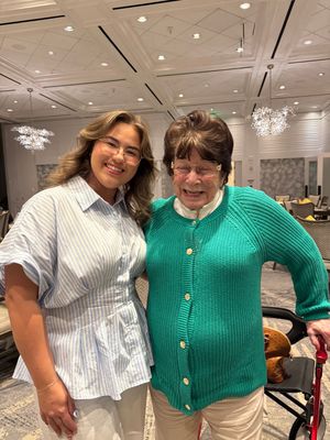 Two women smiling together in an elegant hotel lobby, one in a blue striped shirt and one in a green cardigan.